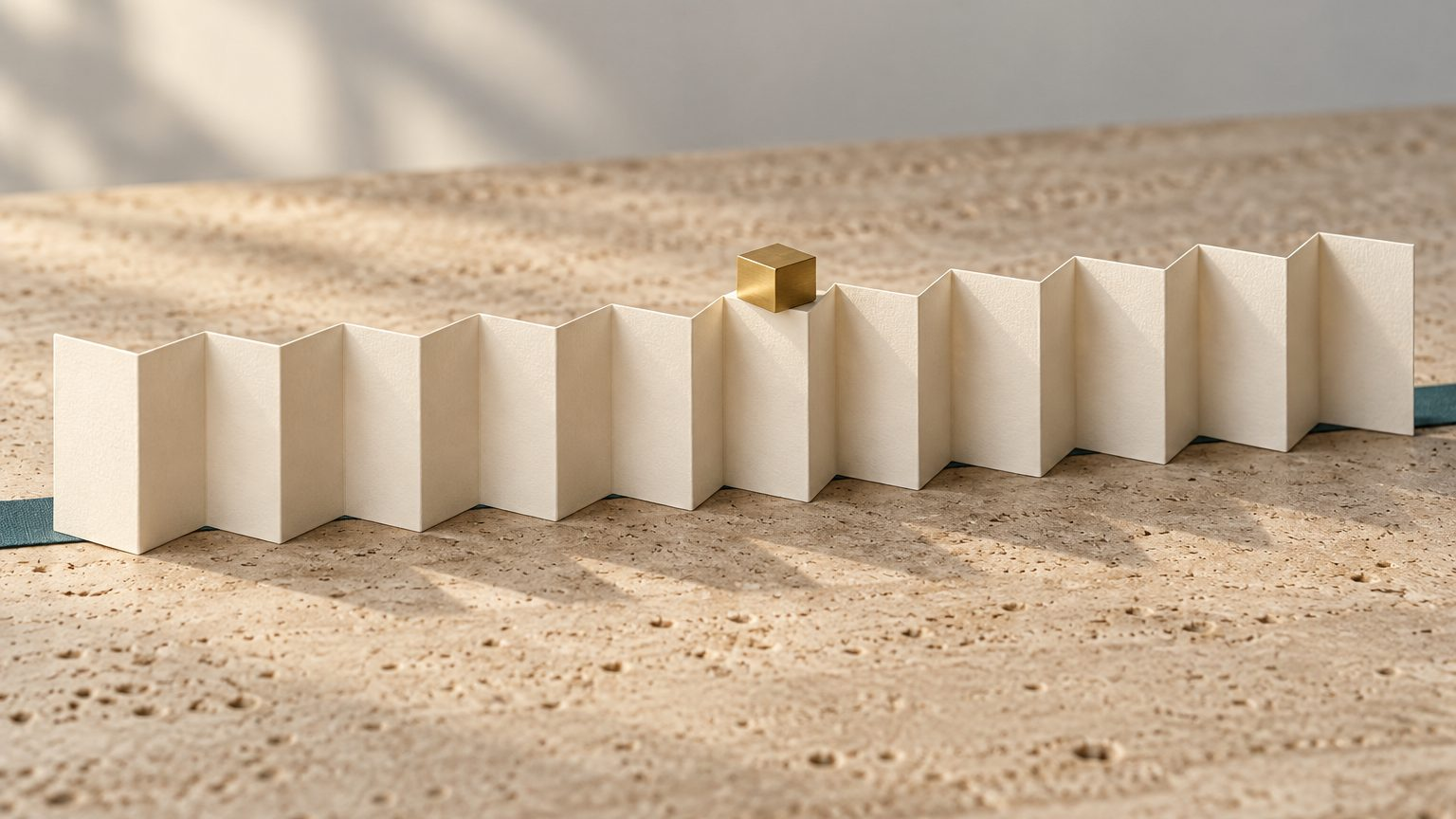 A long strip of cream paper folded into twelve accordion panels on travertine, with a small brass cube resting on the seventh panel